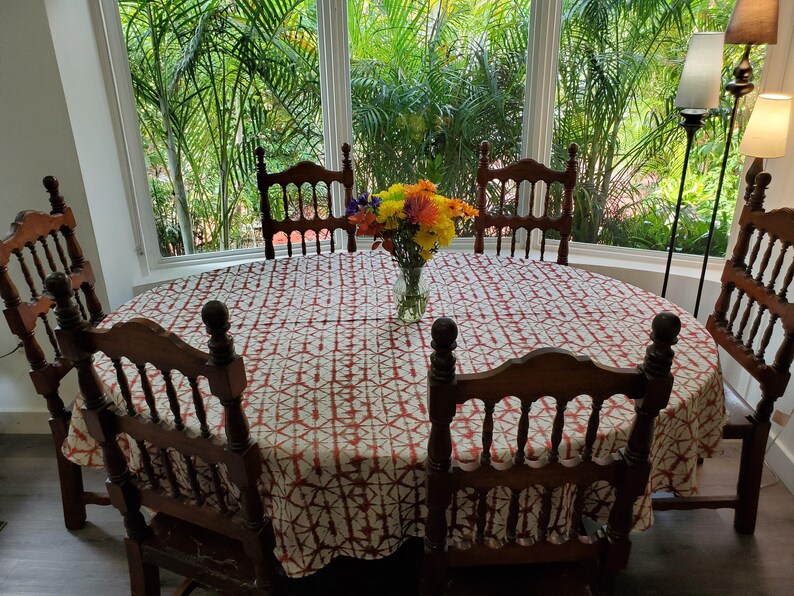 A oval wooden dining table set with six chairs. The table is covered with a Shibori tablecloth in red and white. A vase of colorful flowers sits in the center. The chairs are made of dark wood.