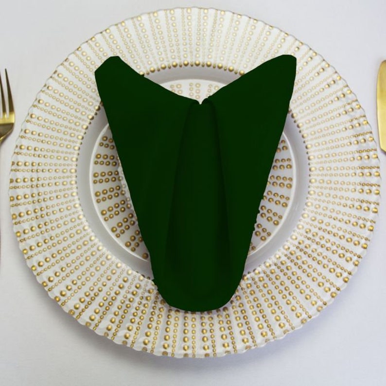 A dark green linen napkin folded into a triangle shape on a white charger plate with a gold beaded design.