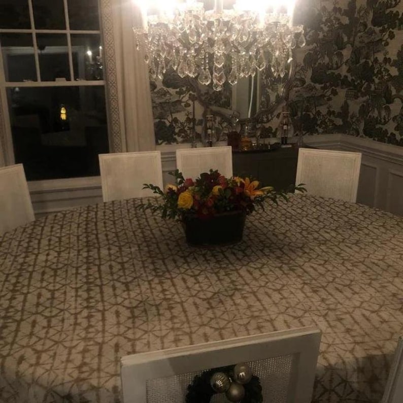 A formal dining room with a large oval  table cloth. It is a Shibori tablecloth. A floral arrangement sits in the center. White chairs surround the table. A crystal chandelier hangs above, and a window is visible in the background.