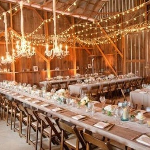 A rustic barn wedding reception venue with long wooden tables, white linens, white burlap table runners and string lights hanging from the ceiling. The tables are set with white plates, silverware, and glasses. There are flowers on the tables and candles in glass jars.