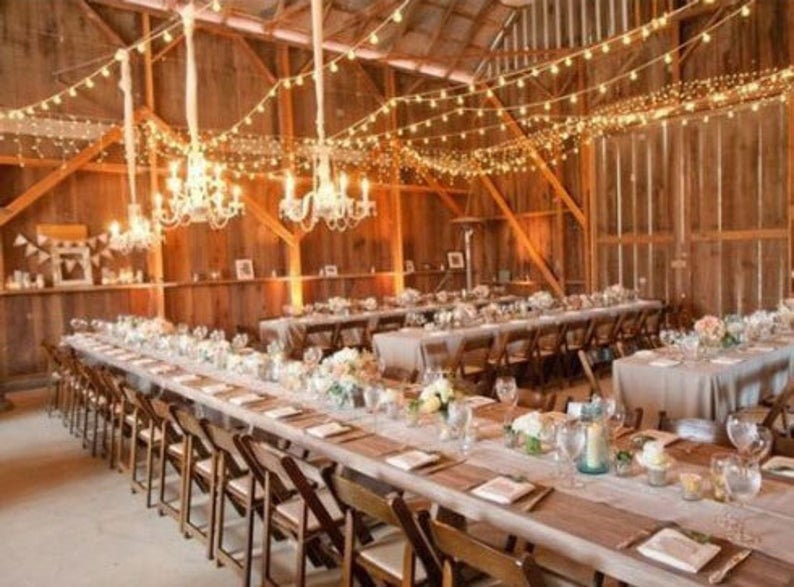 A rustic barn wedding reception venue with long wooden tables, white linens, white burlap table runners and string lights hanging from the ceiling. The tables are set with white plates, silverware, and glasses. There are flowers on the tables and candles in glass jars.