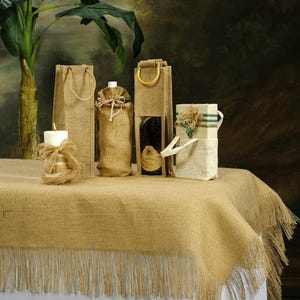 May include: Four burlap gift bags with handles, two with bottles inside, one with a candle, and one with a box. The bags are sitting on a brown burlap table runner with fringe.