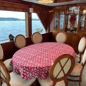 A dining table set on a yacht, featuring a oval table  cloth. It is a  a red and white Shibori tablecloth. The table is surrounded by wooden chairs with cream-colored seats. The background shows a large window with a view of the water.