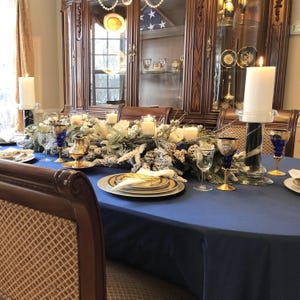 Formal, large tablecloth in blue on an oval table with cotton napkins, fine china, floral arrangements and candles. Very elegant table and dining room.