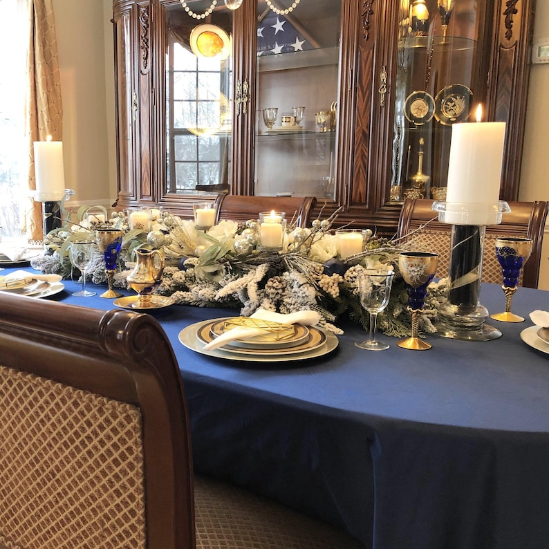 Formal, large tablecloth in blue on an oval table with cotton napkins, fine china, floral arrangements and candles. Very elegant table and dining room.