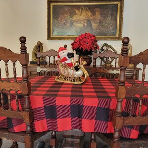 May include: A round wooden dining table with a red and black buffalo plaid tablecloth. There are six wooden chairs around the table, and a small Santa Claus figurine in a sleigh is in the center of the table.