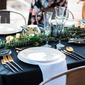 A black tablecloth, white cotton napkin with gold flatware, white plates, and black and gold glasses. A sprig of greenery with white flowers runs down the center of the table.