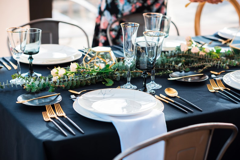 A black tablecloth, white cotton napkin with gold flatware, white plates, and black and gold glasses. A sprig of greenery with white flowers runs down the center of the table.