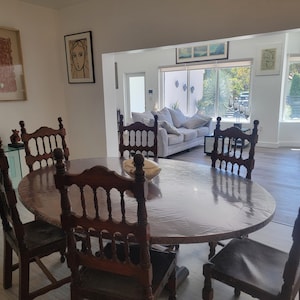 May include: A wooden dining table with six chairs. The table is oval shaped and has a light brown finish. The chairs are also wooden and have a dark brown finish. The table is set with a white tablecloth and a bowl of fruit in the center.