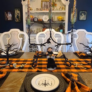 A Halloween table setting with a black and orange buffalo plaid tablecloth and checkered napkins. The table is set with black plates, gold chargers, and black wine glasses. There are also black and white skull decorations on the table.