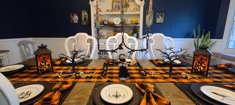 A Halloween table setting with a black and orange buffalo plaid tablecloth and checkered napkins. The table is set with black plates, gold chargers, and black wine glasses. There are also black and white skull decorations on the table.