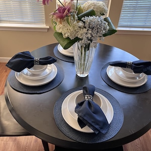 A round table in the dining room of a home with the table set for a meal and black napkins in napkin rings on top of the plates. A large vase of flowers is in the center of the table.