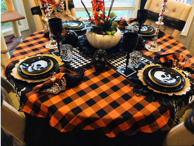 A Halloween table setting with a black and orange checkered tablecloth. The table is set with black plates, gold chargers, and black wine glasses. There are also black and white skull decorations on the table.