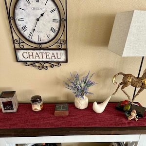 May include: A decorative display featuring a wall clock with the word "Chateau" and a table with various ornaments. The table includes a small vase with lavender, a jar, a white bird figurine, and a lamp with a beige lampshade.