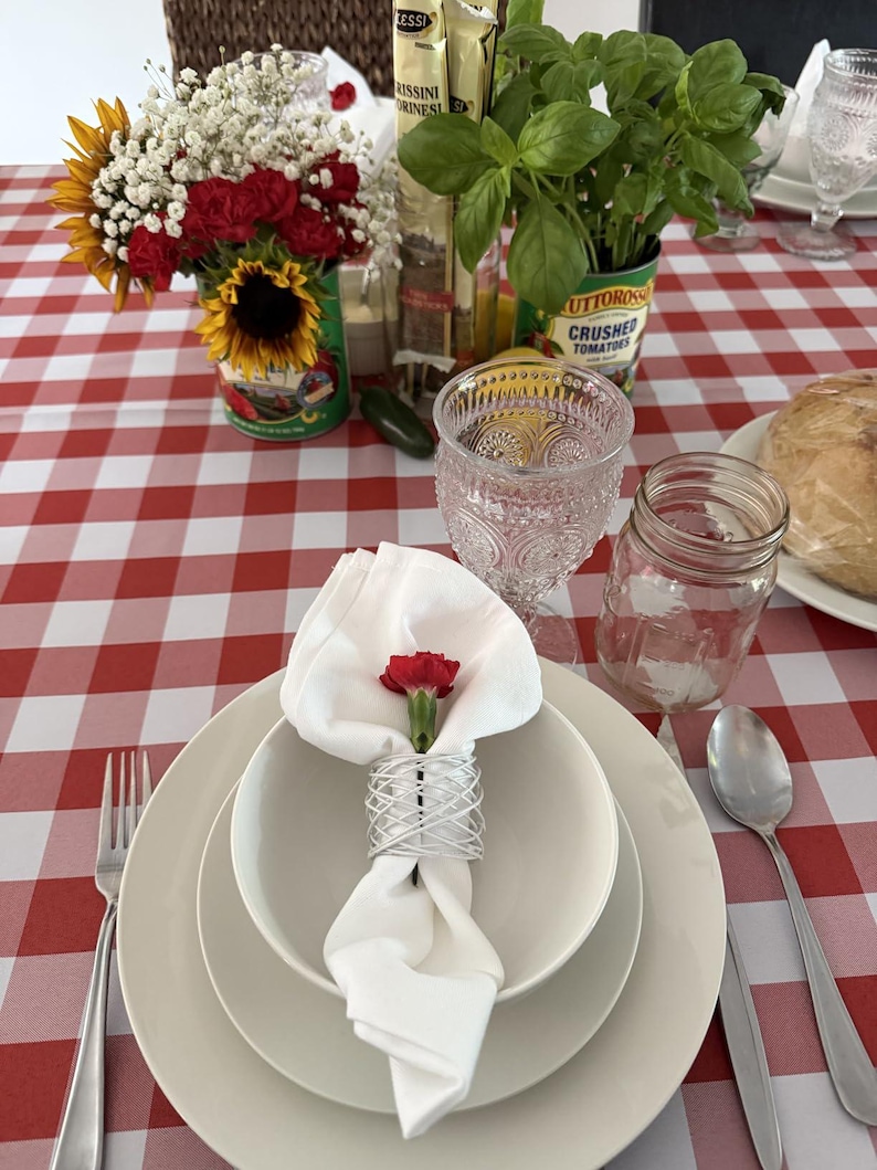 A table with a checkered red and white tablecloth and a napkin on top of the china with a red flower in the dinner napkin.