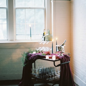 A bar cart with a dark purple velvet table runner draped over it. The cart is filled with glassware, including champagne flutes and wine glasses. There are also candles and a bottle of champagne in an ice bucket.