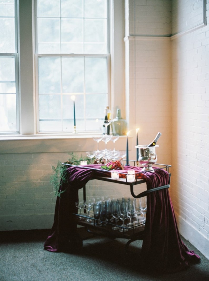 A bar cart with a dark purple velvet table runner draped over it. The cart is filled with glassware, including champagne flutes and wine glasses. There are also candles and a bottle of champagne in an ice bucket.