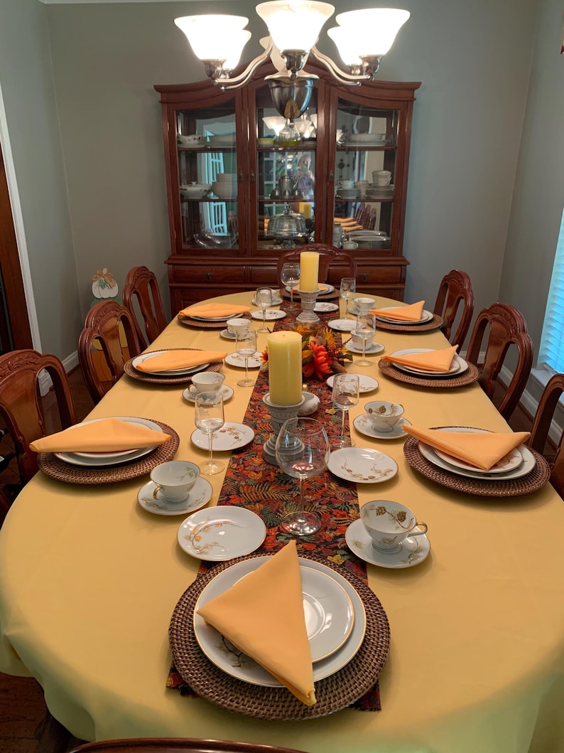 A yellow oval tablecloth, dinner napkins and red table runner in the dining room of a home. China, silverware and glasses as the table is ready for an elegant meal.