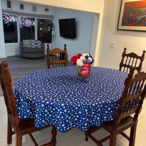 May include: A round dining table covered with a blue tablecloth featuring a white star pattern, surrounded by four dark wood chairs. A centerpiece sits on the table, consisting of red, white, and blue pom-poms and a sign that reads God Bless America.