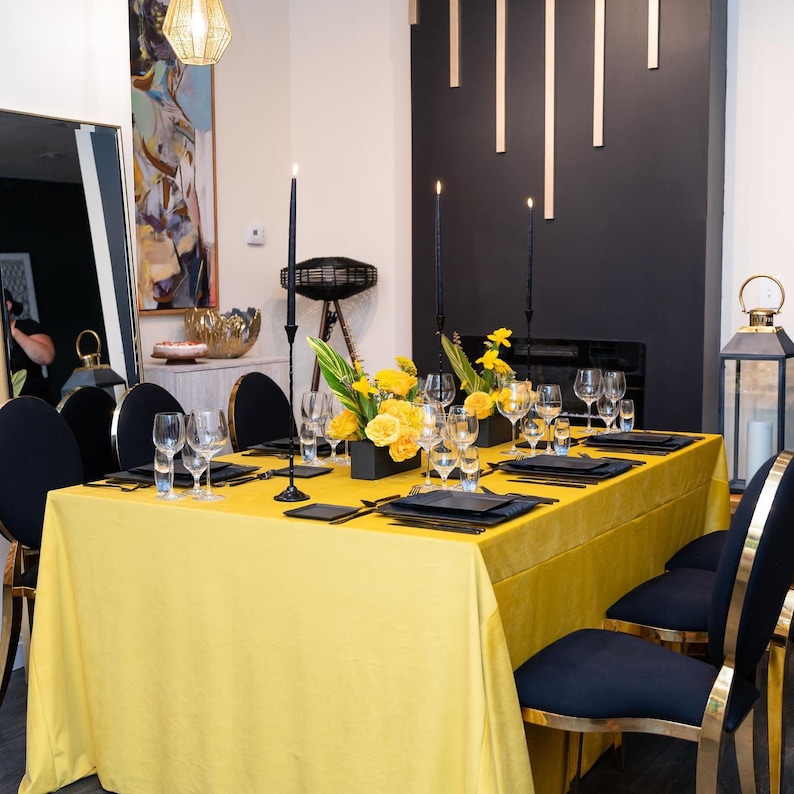 A yellow velvet tablecloth with black candlesticks and a centerpiece of yellow flowers. The table is set with black plates, black velvet napkins, and clear glasses. There are two black chairs with gold accents in the foreground.
