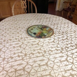 A champaign oval tablecloth with a geometric pattern covers a table. A decorative glass cutting board with a fruit and wine design sits on top. A wooden chair is in the background.