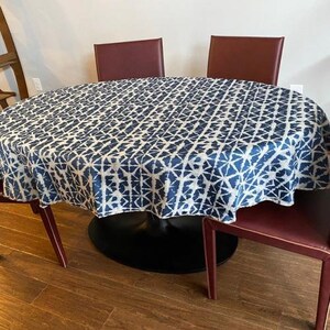 May include: A round dining table covered with a blue and white patterned tablecloth. The table is surrounded by burgundy chairs. The tablecloth has a geometric design. The table has a black base and wooden floor.