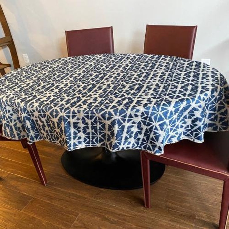 May include: A round dining table covered with a blue and white patterned tablecloth. The table is surrounded by burgundy chairs. The tablecloth has a geometric design. The table has a black base and wooden floor.