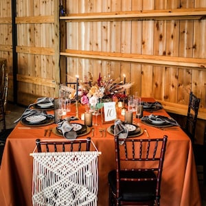 May include: A rustic wedding table setting with a burnt orange tablecloth, black and gray place settings, and a centerpiece of fall flowers. The table is set with candles and a gold table number card that says "Table 10".