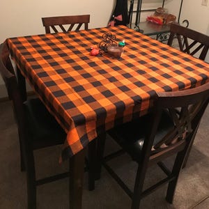A black and orange Buffalo plaid tablecloth on a wooden table with four chairs. The tablecloth is made of a soft, woven fabric and has a classic buffalo plaid pattern.
