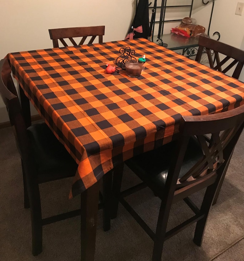 A black and orange Buffalo plaid tablecloth on a wooden table with four chairs. The tablecloth is made of a soft, woven fabric and has a classic buffalo plaid pattern.
