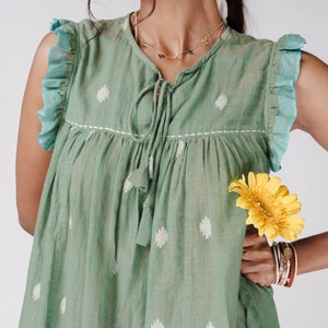 May include: A green sleeveless dress with white polka dots and a tie at the neckline. The dress has a ruffled trim on the sleeves. A yellow flower is held in the right hand of the person wearing the dress.