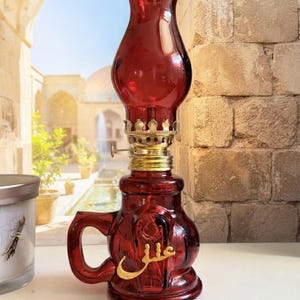 May include: A deep red glass oil lamp featuring a bulbous base with a handle and a matching chimney top. The base is adorned with gold-colored Arabic calligraphy. A metallic gold burner assembly connects the glass components, featuring a small adjustment knob.