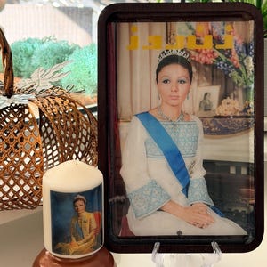 May include: A rectangular dark wood tray featuring a portrait of Empress Farah Pahlavi in a white gown with a blue sash and tiara, with yellow Persian text at the top. Beside it sits a white pillar candle displaying the same portrait.