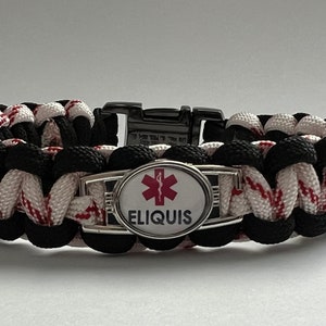 May include: A medical alert bracelet featuring a woven paracord band in black and white with red accents. The center includes a silver-toned oval plate displaying a red Star of Life symbol above the black text ELIQUIS. It has a black plastic buckle.