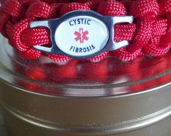 May include: A red paracord bracelet featuring a central silver-toned metal plate with an oval white inlay. The inlay displays the text CYSTIC FIBROSIS in black lettering above and below a red medical Star of Life symbol with a central serpent staff.