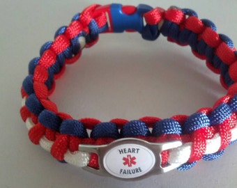 May include: A paracord medical alert bracelet featuring a woven pattern of red, white, and blue nylon cord. The bracelet includes a blue plastic side-release buckle and a central silver-tone metal plate with a red star of life and the text HEART FAILURE.