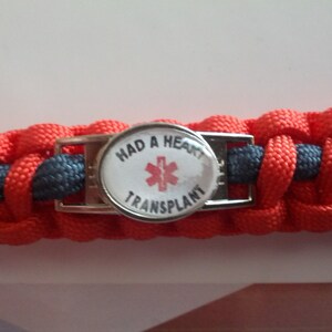 Puede incluir: Una pulsera de paracord roja y gris con una hebilla plateada y un bot&oacute;n redondo blanco que dice "Had a Heart Transplant" con un s&iacute;mbolo m&eacute;dico rojo.