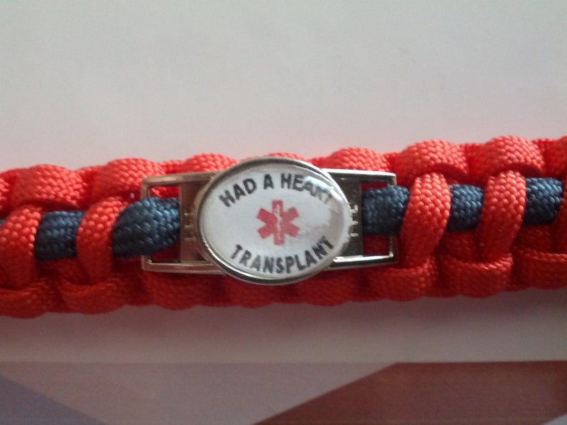 Puede incluir: Una pulsera de paracord roja y gris con una hebilla plateada y un bot&oacute;n redondo blanco que dice "Had a Heart Transplant" con un s&iacute;mbolo m&eacute;dico rojo.