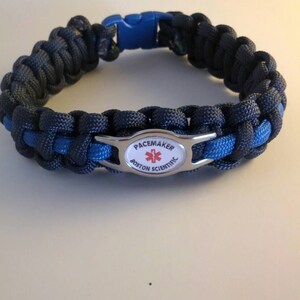 May include: A paracord bracelet woven from dark navy and bright blue cord, featuring a blue plastic side-release buckle and a central silver-toned metal plate with a white inlay. The plate displays a red medical star and the text PACEMAKER BOSTON SCIENTIFIC.