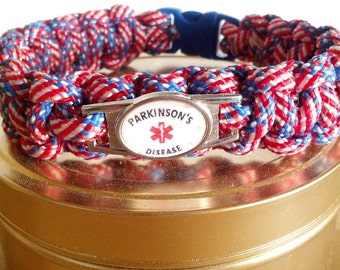 May include: A medical alert bracelet made of braided red, white, and blue paracord. The bracelet features a central silver-toned metal plate with an oval white inlay displaying the text PARKINSON'S DISEASE and a red Star of Life symbol.