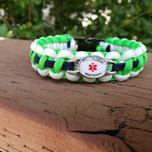 May include: A paracord bracelet woven with bright green, white, and dark blue nylon cord. The bracelet features a central silver-toned oval medical alert charm with a red star of life symbol and the text PACEMAKER MEDTRONIC printed on it.