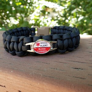 May include: A black paracord bracelet featuring a central oval metal charm with a red medical alert symbol and the text MEDICAL ALERT BLOOD THINNERS. The bracelet includes a tan plastic side-release buckle for closure and a woven cord design.