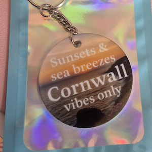 May include: A round keychain with a silver ring and chain. The keychain features a circular pendant with the words "Sunsets & sea breezes Cornwall vibes only" over a coastal image. It is packaged in a holographic pouch.