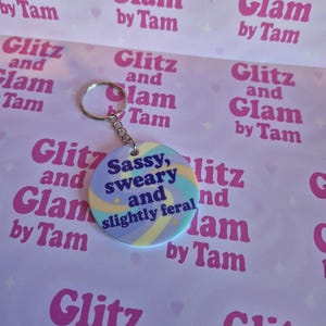 Puede incluir: Un llavero colorido con un dije redondo que dice "Sassy, sweary and slightly feral". El dije está en una cadena plateada con un anillo plateado. El fondo es de color morado claro con un patrón repetido de corazones rosas y estrellas. El texto "Glitz and Glam by Tam" se repite en todo el fondo.