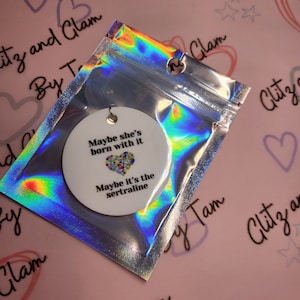 May include: A white round ornament with the text "Maybe she's born with it. Maybe it's the sertraline." and a colorful heart graphic. The ornament is in a silver iridescent bag.