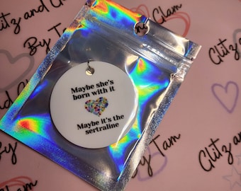 May include: A white round ornament with the text "Maybe she's born with it. Maybe it's the sertraline." and a colorful heart graphic. The ornament is in a silver iridescent bag.