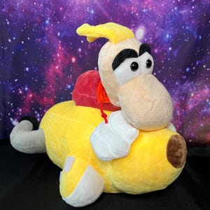 May include: A plush toy character featuring a yellow body, a red hooded garment, and a large head with white skin, black eyebrows, and black eyes. The figure has white hands, a brown nose, and a grey tail, presented in a soft, stuffed construction.