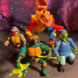 Puede incluir: Un grupo de figuras de acción de las Tortugas Ninja. Las figuras están en varias poses y colores, incluyendo verde, naranja, morado y azul. Una figura es un gran monstruo naranja con detalles rojos y amarillos.