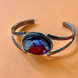 May include: Silver-toned cuff bracelet with an oval, dichroic glass centerpiece. The glass features a mix of red, blue, and silver colors. The bracelet has an open design, allowing for easy wear.