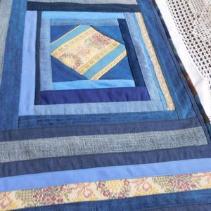 Könnte beinhalten: Ein rechteckiger Denim-Quilt mit einem zentralen Rautenmuster aus gelbem und floralem Stoff. Der Quilt weist verschiedene Blautöne von Denim-Streifen auf, die ein Patchwork-Design erzeugen. Der Quilt ist etwa 61 cm lang und 30 cm breit.